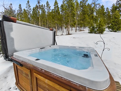 An outdoor hot tub filled with water sits on a snow-covered deck, surrounded by a scenic pine forest.