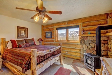 A rustic bedroom with log walls, a large bed with patterned bedding, a stone fireplace, and a window showing a snowy mountain view. A ceiling fan and decorative art pieces are also present.