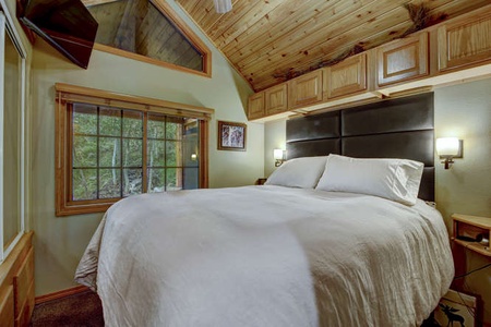 A cozy bedroom with a large bed, white bedding, wood-paneled ceiling, built-in cabinets, a window, wall-mounted TV, and bedside lights.