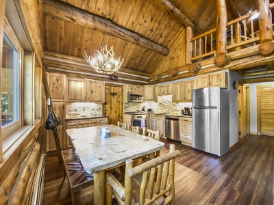 A rustic kitchen with wooden cabinetry, a large stainless steel refrigerator, an antler chandelier, and a wooden dining table surrounded by matching chairs. The space features a loft area above.