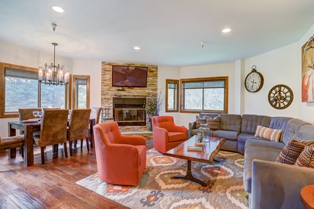 A spacious living and dining area with a stone fireplace, grey sectional sofa, two orange chairs, a wooden dining table with eight chairs, and large windows with outdoor views.