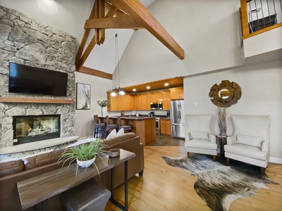 Spacious living area featuring vaulted ceilings, exposed wooden beams, stone fireplace with mounted TV, wooden dining set, modern kitchen, two white armchairs, and cowhide rug over hardwood floors.