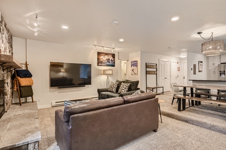 A modern living room with a wall-mounted TV, a stone fireplace, several seats, and a dining area with a bench and chairs. The room is well-lit, and there are pictures and decor on white walls.