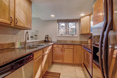 A small kitchen with wooden cabinets, granite countertops, stainless steel appliances, a double sink, dishwasher, and a window above the counter. A coffee maker and soap dispenser are on the counter.