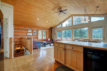 Spacious cabin living area with wooden interiors, a kitchen island, seating area with sofas, large windows offering forest views, and a ceiling fan.