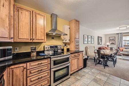 A contemporary kitchen with wooden cabinets, a stainless steel stove, and a range hood, opens into a dining area and living room with a sofa and wall art.