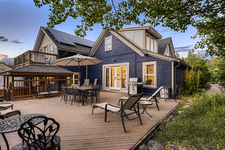 Backyard view of a house with blue siding, featuring a wooden deck, outdoor dining furniture, lounge chairs, an umbrella, a barbecue grill, and adjacent greenery.