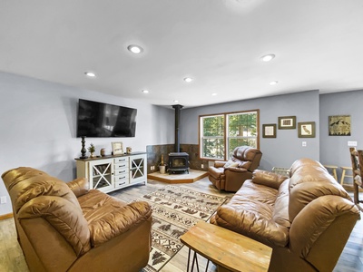 A cozy living room with brown leather recliners, a wooden coffee table, a flat-screen TV, a wood-burning stove, and a large window letting in natural light. Decor includes a patterned rug and wall art.