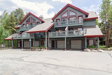 A multi-unit building with three floors and garages on the ground level. The structure has gray siding with red trim and is surrounded by trees. The scene is set on a clear day with blue skies.