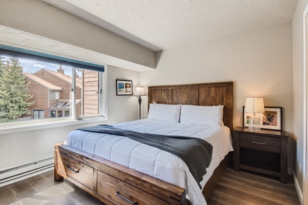 A cozy bedroom with a double bed featuring white sheets and a gray throw blanket, a wooden headboard, and matching bedside table with a lamp. Large window with a view of neighboring buildings and trees.