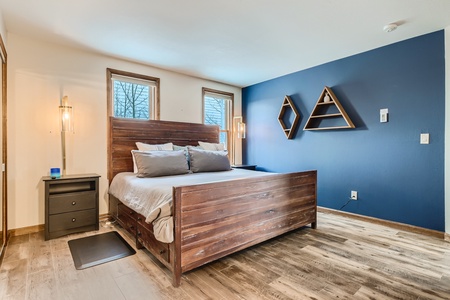 A bedroom with a wooden bed frame, gray bedding, two side tables with lamps, wooden floors, blue accent wall, and geometric wall shelves.