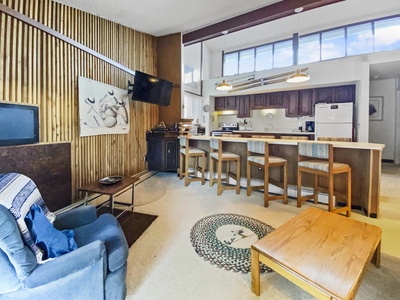 A cozy living space with a blue armchair, wooden coffee table, breakfast bar with stools, and a mounted TV on a wooden panel wall. Kitchen in the background with white appliances and high windows above.