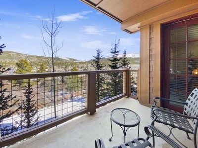 A balcony with metal chairs and a small table, overlooking a scenic mountain landscape with evergreen trees and a partly cloudy sky.