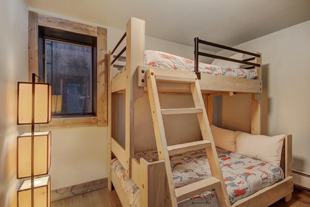 A small bedroom featuring a wooden bunk bed with colorful bedding, a window on the left wall, and a tall lamp with three square shades.