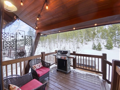 Covered wooden deck with string lights, a black grill, two black wicker chairs with red cushions, and a small glass-top table. Snow-covered landscape with trees is visible in the background.