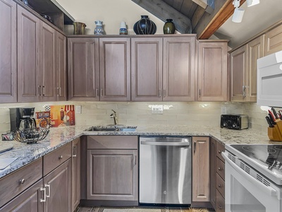 Modern kitchen with wooden cabinets, stainless steel dishwasher, electric stove, microwave, granite countertops, and various kitchen items including a coffee maker, knife set, and decorative jars.