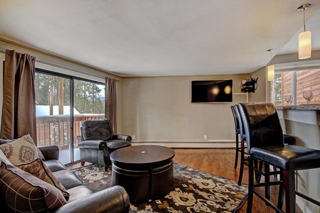 A cozy living room features a leather couch, armchairs, a TV on the wall, and a small dining area with high stools. Large windows and a sliding glass door lead to a balcony with a view.