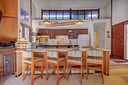 A kitchen area with wooden cabinetry, a white fridge, and a stove. A counter with four striped upholstered bar stools faces the kitchen. Overhead, skis are mounted as decoration.