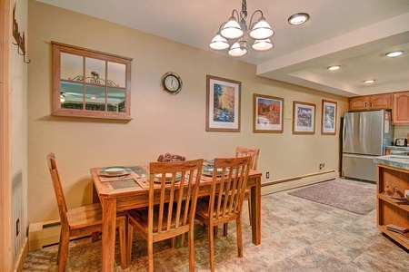 Dining area with a wooden table and chairs, chandelier, wall art, a clock, and a window. Kitchen area with stainless steel refrigerator and wooden cabinets is visible in the background.