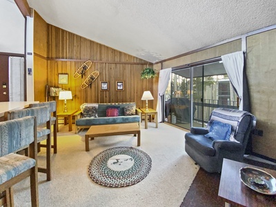 Cozy living room with a sofa, armchair, wooden coffee table, barstools, and a balcony. The room is decorated with framed pictures, wall decor, and indoor plants.