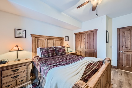 A cozy bedroom with a rustic wooden bed frame, plaid bedding, wooden nightstands, a ceiling fan, artwork on the wall, and a closed wooden closet door. The room has a warm, inviting atmosphere.