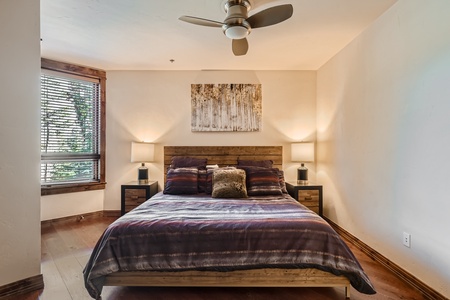 A cozy bedroom with a wooden bed, nightstands, lamps, a ceiling fan, a window with blinds, and a nature-themed artwork above the bed.