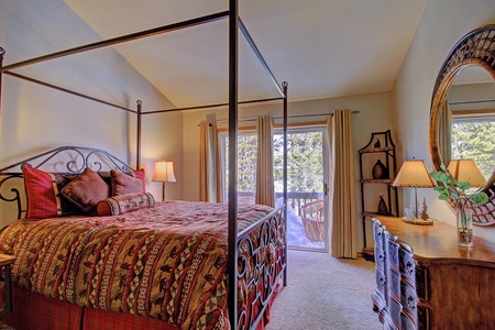 A bedroom with a four-poster bed, patterned bedding, and multiple pillows. It has a balcony, a dresser with two lamps, a mirror, and a round wicker chair outside. Curtains cover a glass door.