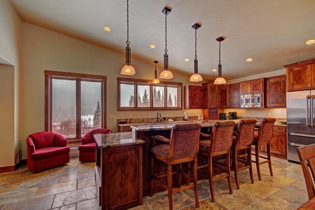 A spacious kitchen-dining area features wooden cabinetry, granite countertops, bar seating with high chairs, pendant lights, stainless steel appliances, and two red armchairs near large windows.