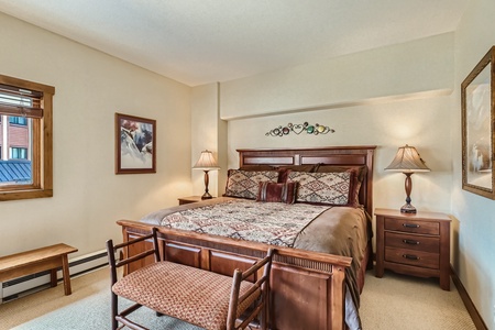 A cozy bedroom with a wooden bed, patterned bedspread, two bedside tables with lamps, a bench at the foot of the bed, and wall art. The room has beige walls and a window on the left side.