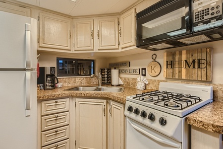 A small kitchen features a white refrigerator, gas stove, microwave, sink, and coffee maker. Decorative signs on the wall read "HOME" and "JOY-LIFE." Cabinets and countertops are light brown.