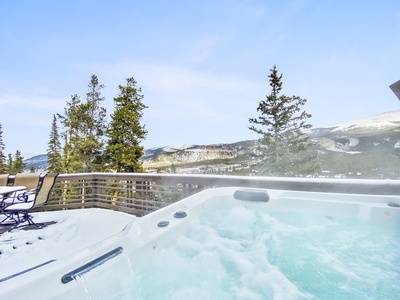 A hot tub on a deck surrounded by snow-covered trees and mountains on a clear day.