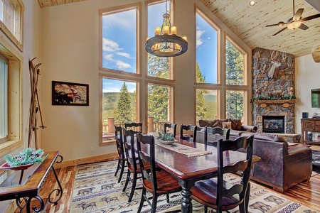 A spacious dining and living area with large windows, a wooden dining table, black chairs, a stone fireplace, and an antler chandelier. The room features rustic decor and a mountain view outside.