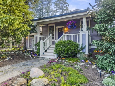 A small house with a front porch surrounded by various plants, flowers, and shrubs. The porch has stairs leading up to a door and a hanging flower basket on the side.