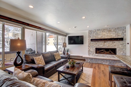 A modern living room features leather sofas, a stone fireplace, a mounted TV, large windows, and wooden flooring.