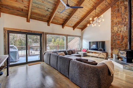 A cozy living room with a vaulted wood ceiling, large sectional sofa, modern chandelier, flat-screen TV, stone fireplace, and sliding glass doors leading to a deck with a scenic outdoor view.