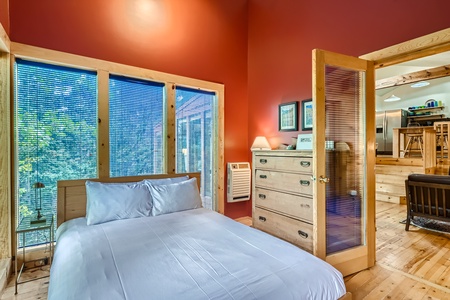 A cozy bedroom with red walls features a double bed, blue bedding, a wooden dresser, and large windows with blue blinds and a view of trees outside. An air conditioning unit is mounted on the wall.