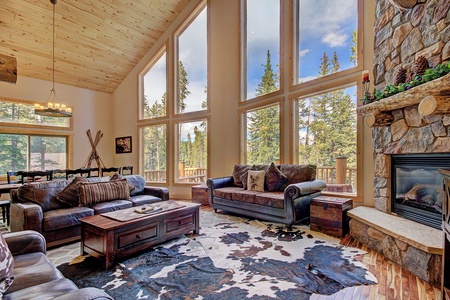 A spacious living room with large windows showcasing a forest view. It features leather sofas, a cowhide rug, a wooden coffee table, a stone fireplace, and a high, wood-paneled ceiling.