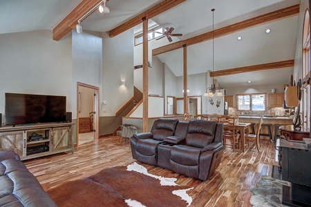 A spacious living room with high ceilings, exposed wooden beams, leather recliners, a large TV, and an open plan leading to the kitchen and dining area. A cowhide rug is on the wooden floor.