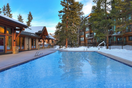 Outdoor swimming pool bordered by a snowy landscape and surrounded by buildings and trees under a clear blue sky.