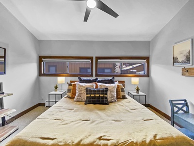 A bedroom with a bed, three pillows, two side tables with lamps, gray walls, three narrow windows, a ceiling fan, and a chair.