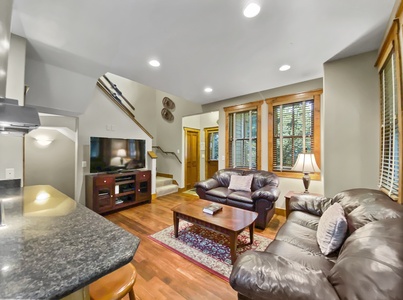 A cozy living room with leather sofas, a wooden coffee table, and a flatscreen TV on a wooden stand. To the right, there is a window and a lamp, and stairs leading up in the background.