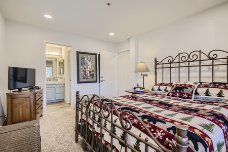 A bedroom with a metal bed frame, festive bedding, a nightstand with a lamp, a TV on a wooden dresser, and an open door leading to a bathroom.