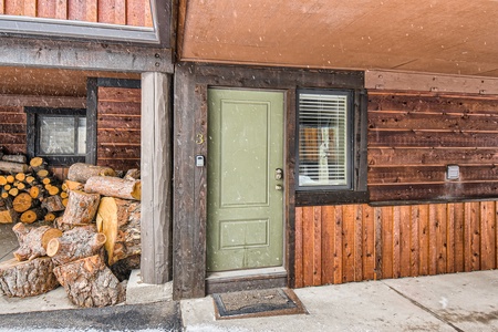 An exterior view of a wooden house entrance with a green door marked "3," a window with blinds, a pile of chopped logs, and falling snow.