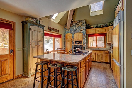 Rustic kitchen with wooden cabinets and island, granite countertops, a stone backsplash around the stove, and skylights. Four wooden bar stools are placed around the island.