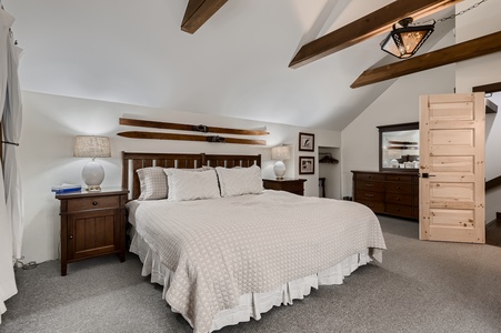 A bedroom with a large bed, wooden beams on the ceiling, a dresser, and a door leading to another room. The bed has a white quilt, and there are two bedside tables with lamps.