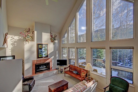 Spacious living room with large floor-to-ceiling windows, high ceilings, and natural light. Furnished with a fireplace, various seating, a television, and nature-themed decor.