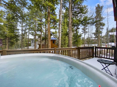 A hot tub on a wooden deck is surrounded by tall trees under a partly cloudy sky. A small building is visible in the background.