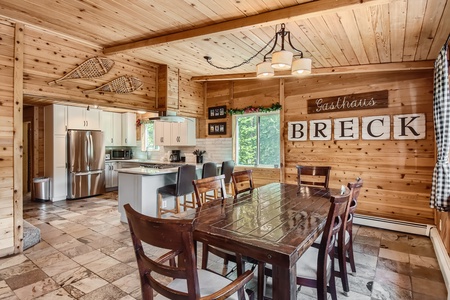 A rustic wooden dining room with a polished table, chairs, a modern kitchen with stainless steel appliances, and a sign on the wall that reads "Gästehaus BRECK." Snowshoes and plants adorn the space.