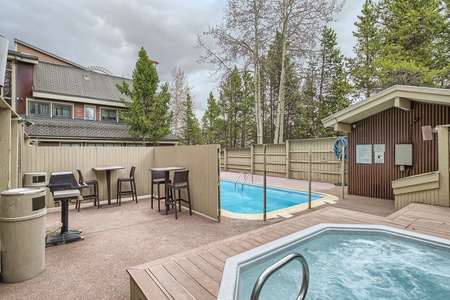 A small outdoor pool and hot tub are enclosed by a wooden fence, with trees and a building in the background. Adjacent are two high-top tables with black chairs and a grill.