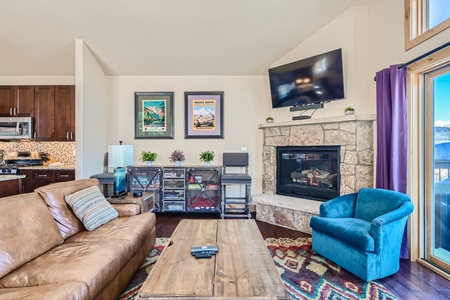 A cozy living room with a brown leather sofa, a blue armchair, a wooden coffee table, a stone fireplace, and a mounted TV. Two framed artworks hang on the wall above a shelf with decor items.